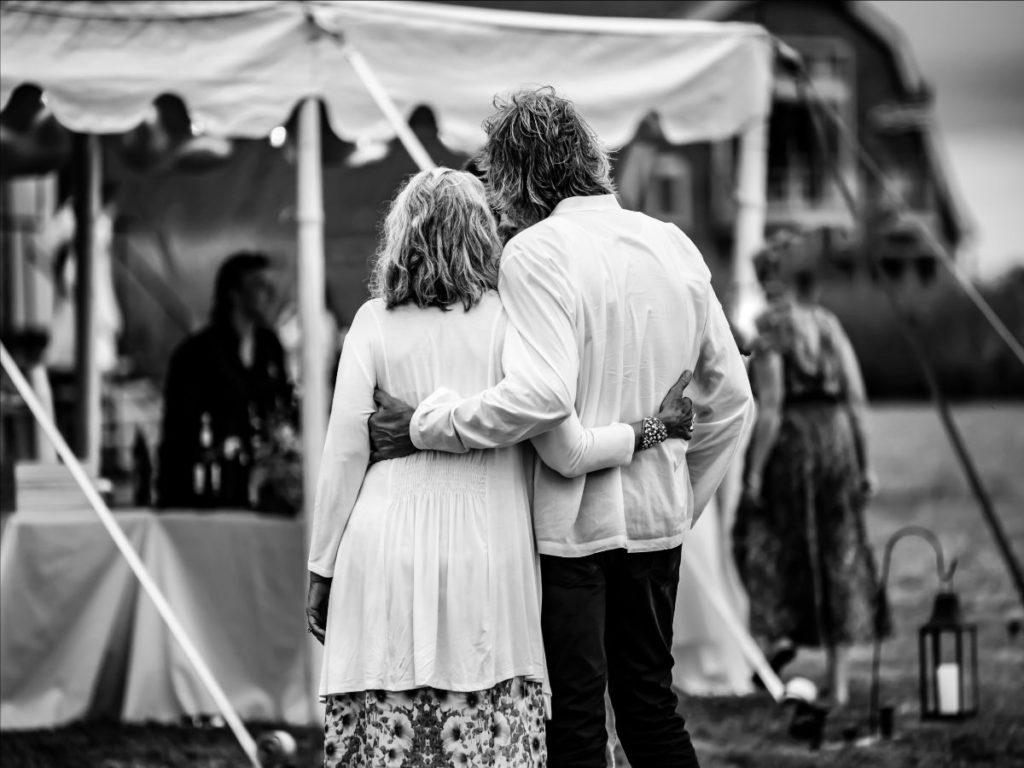 Two people appear at center of a black & white image. They are facing away from the camera and towards the tent, with their arms around one another's waists.
