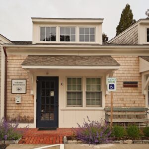 A photograph of an office frontage on a cedar-shingled building. To the left of the door is a white sign with green logo & text for Sakonnet Preservation. In front of the brick walkway are various spring flowers in bloom.